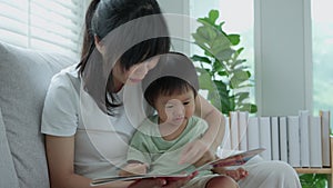 Happy Asian mother relax and read book with baby time together at home. parent sit on sofa with daughter and reading a story.