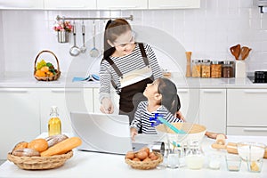 Happy Asian mother teaching her young daughter to bread baking in white modern kitchen by looking at recipe online from computer