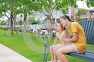 Happy asian mum and daughter relaxing sitting on bench in garden outdoors. Mother pointing something with child girl looking in