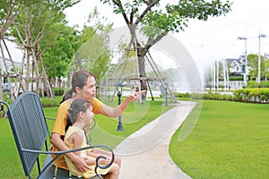 Happy asian mum and daughter relaxing sitting on bench in garden outdoors. Mother pointing something with child girl looking in