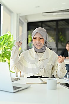 Happy Asian Muslim female office worker hands up, celebrating her success, project approved