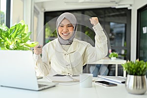 Happy Asian Muslim female office worker hands up, celebrating her success, receiving good news