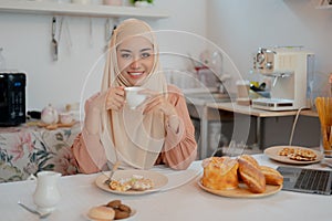 A happy Asian Muslim woman is sipping hot tea and having breakfast at a table in the kitchen