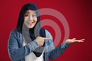 Happy Asian muslim woman smiling at camera while presenting and pointing something with open palm hand, holding product in copy