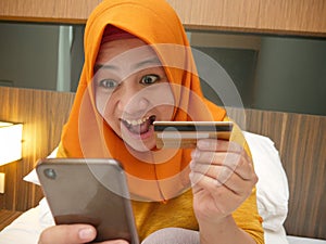 Happy Asian muslim woman wearing hijab smiling and laughing, when making purchase on online shop with her phone and credit card