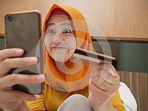 Happy Asian muslim woman wearing hijab smiling and laughing, when making purchase on online shop with her phone and credit card