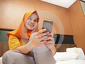 Happy Asian muslim woman wearing hijab smiling when reading text message or chat on her phone while lying on bed