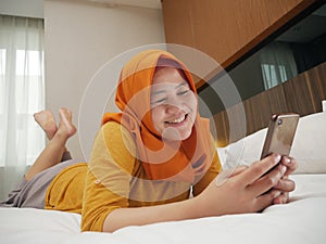 Happy Asian muslim woman wearing hijab smiling when reading text message or chat on her phone while lying on bed