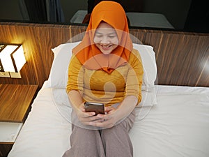 Happy Asian muslim woman wearing hijab smiling when reading text message or chat on her phone while lying on bed