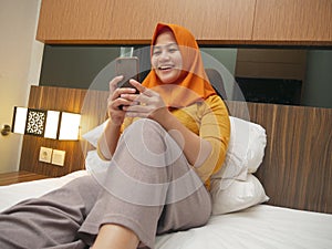 Happy Asian muslim woman wearing hijab smiling when reading text message or chat on her phone while lying on bed