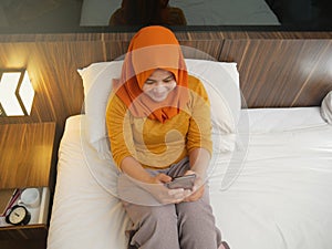 Happy Asian muslim woman wearing hijab smiling when reading text message or chat on her phone while lying on bed