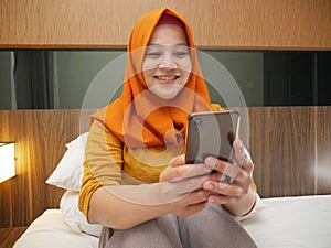 Happy Asian muslim woman wearing hijab smiling when reading text message or chat on her phone while lying on bed