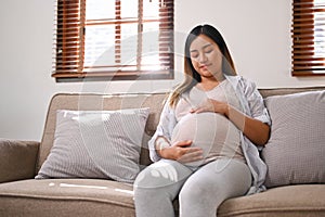 Happy Asian pregnant woman in comfortable clothes relaxes on the sofa in her living room