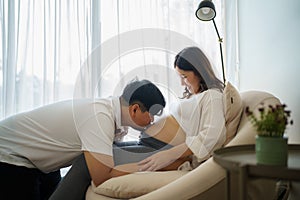 Happy Asian pregnant woman with husband expecting baby at home and husband try to kiss the baby through belly