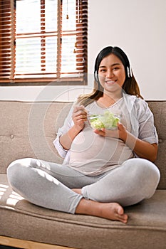 Happy Asian pregnant woman listening to music through her headphones while eating salad
