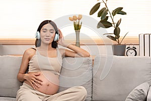 Happy Asian pregnant woman sitting on the bed listen to music in headphones