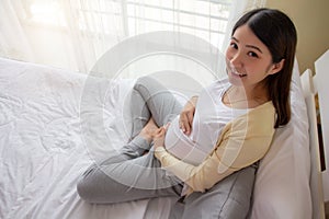Happy Asian pregnant woman sitting on bed touching belly with care at home