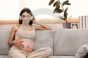 Happy Asian pregnant woman sitting on the couch listen to music in headphones