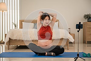 Happy Asian pregnant woman in sportswear sitting on yoga mat stretching warm up and exercise from looking online class by