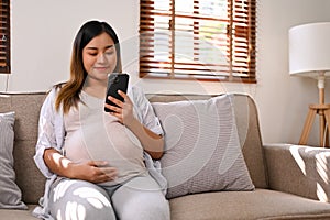 Happy Asian pregnant woman using her phone while relaxing on sofa in her living room