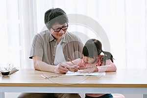 Happy Asian preschool daughter and father in glasses drawing colorful pencils while sit on desk in living room at home. Parents