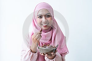 Happy Asian 30s Muslim woman wearing traditional clothes with hijab, holding a bowl of dry dates fruit in hands, smiling at camera