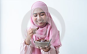 Happy Asian 30s Muslim woman wearing traditional clothes with hijab, holding a bowl of dry dates fruit, smiling looking at fruit