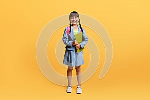 Happy asian schoolgirl with backpack and exercisebook posing on yellow