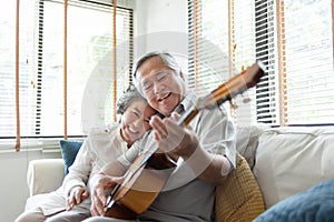 Happy Asian senior Couple enjoying singing and playing acoustic guitar together on sofa at home. Joyful Grandfather and