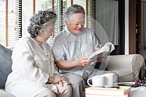 Happy Asian senior couple in love sitting on sofa reading a book together at home