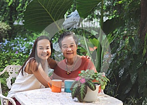 Happy Asian senior woman and her daughter  sitting together at white table in beautiful garden, daughter hugging her mother