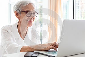 Happy asian senior woman working,surfing the internet with laptop computer at table in home,smiling elderly people in glasses with