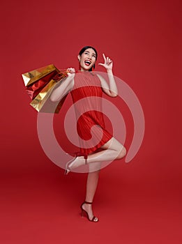 Happy Asian shopaholic woman wearing red dress holding shopping bag isolated on red background. Happy Chinese new year Shopping