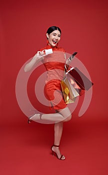 Happy Asian shopper woman wearing traditional cheongsam qipao dress holding credit card and shopping bag isolated on red