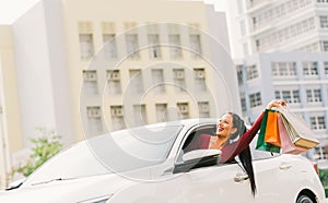Happy Asian tourist woman raise shopping bags on modern white car, look up at copy space. Shopaholic consumerism activity