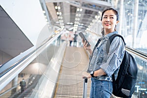 Happy Asian woman with backpack and luggage standing on escalator traveler at international airport. She was using her mobile