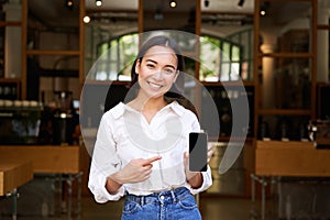 Happy asian woman, cafe manager standing in front of restaurant, pointing finger at smartphone app, showing mobile