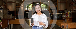 Happy asian woman, cafe manager standing in front of restaurant, pointing finger at smartphone app, showing mobile