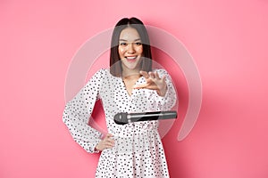 Happy asian woman drop the microphone, smiling and looking confident at camera, standing over pink background