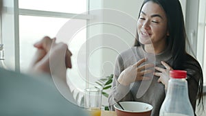Happy Asian woman eating cereal for breakfast and talking to husband smiling in kitchen at home