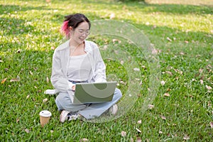 A happy Asian woman in glasses and casual wear sits on the grass in a park working on her laptop