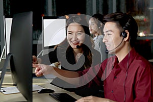 Happy Asian woman headphones work night shift at call center customer care service desk with colleagues, point at computer while