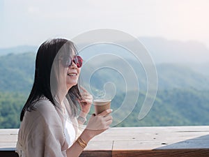 Happy asian woman holding and drinking hot coffee.Smile tourist girl sitting at balcony on mountains and green nature background