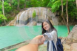 Happy Asian woman holding hand of her boyfriend in honeymoon at Erawan waterfall with trees in national park, tropical forest