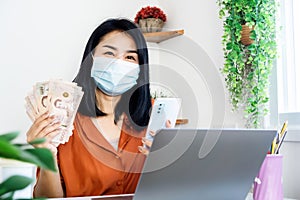 happy Asian woman holding Thai banknote and mobile phone make money online during self-quarantine at home