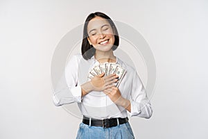 Happy asian woman hugging money dollars and smiling satisfied, standing over white background