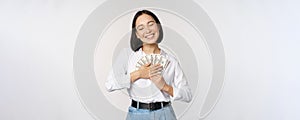 Happy asian woman hugging money dollars and smiling satisfied, standing over white background