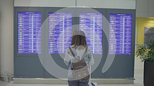 Happy Asian woman looking at information board checking her flight with luggage in terminal hall at the departure gate.