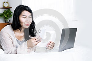 Happy Asian woman paying bills online from home hand holding smart phone scanning qr code