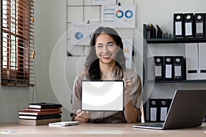 Happy asian woman showing tablet computer with blank screen while sitting at office, mockup for advertising.